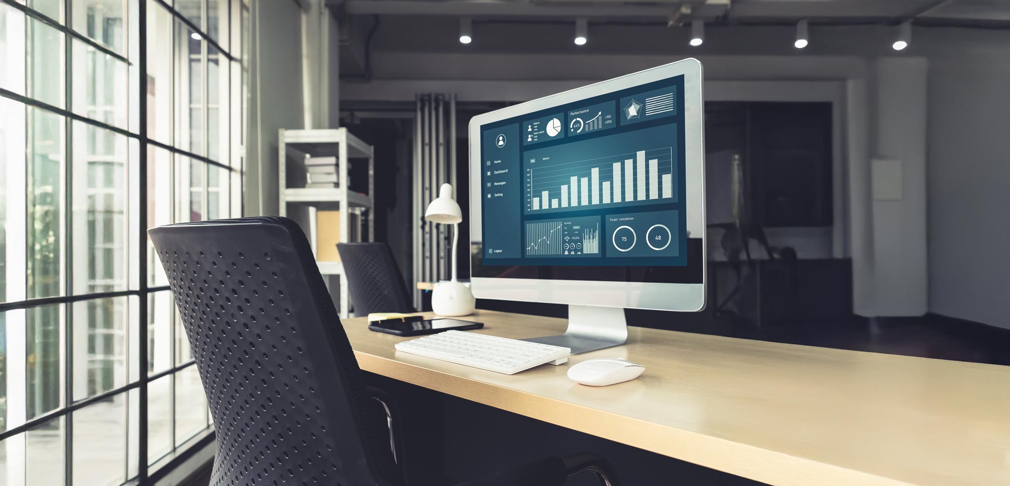 Ein Laptop steht auf einem weißen Schreibtisch in einem hellen, modernen Büroraum und zeigt ein Finanz-Dashboard mit verschiedenen Diagrammen, Balkenstatistiken, einem Kreisdiagramm und Liniengrafen zur Datenanalyse. Im Hintergrund ist ein großer Monitor sichtbar, auf dem eine Website mit Bildern angezeigt wird, sowie eine kleine Topfpflanze, eine Kaffeetasse und weitere Büroausstattung. Der Raum wird durch natürliches Licht erhellt, das durch große Fenster einfällt, und im Hintergrund ist eine Küchenzeile erkennbar.