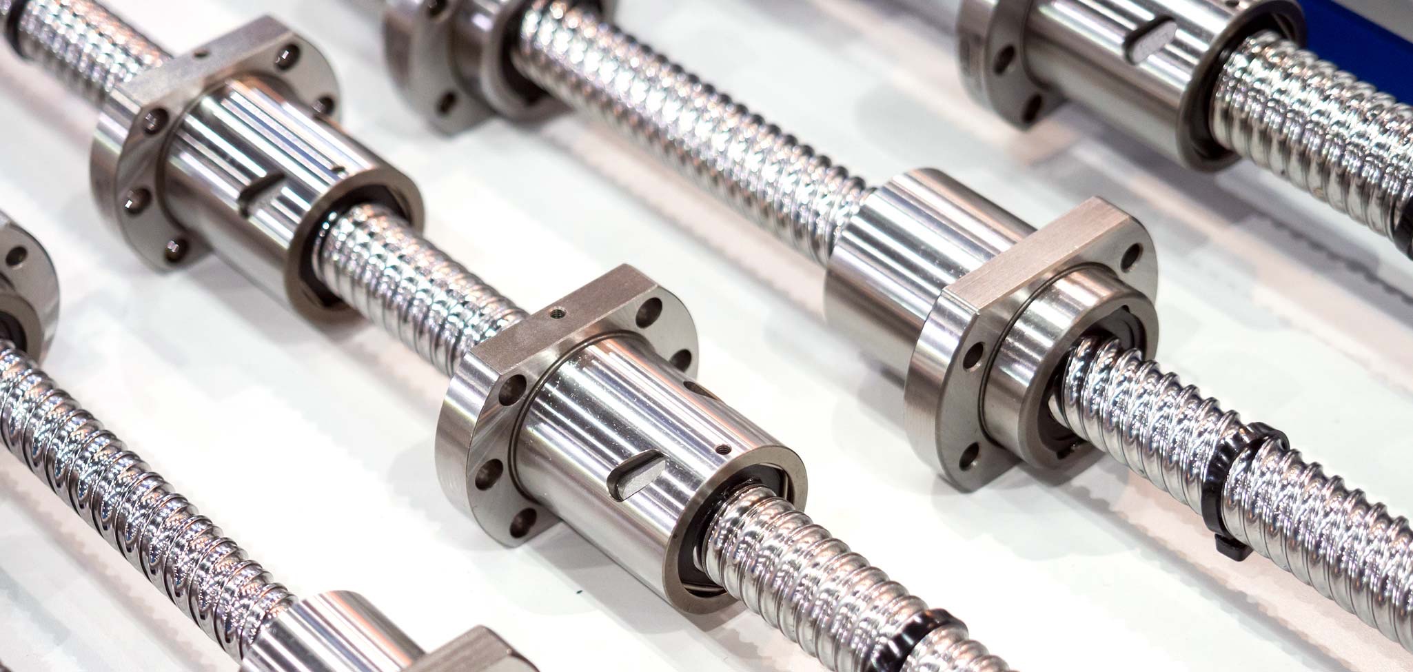 Das Bild zeigt mehrere glänzende Kugelumlaufspindeln aus Metall, die parallel nebeneinander auf einer weißen Fläche angeordnet sind. Die Spindeln haben spiralförmige Gewindegänge und sind mit zylindrischen Kugelumlaufmutter-Bauteilen sowie schwarzen Befestigungsflanschen ausgestattet. Die Anordnung und Beleuchtung betonen die präzise, technische Ausführung der Komponenten.