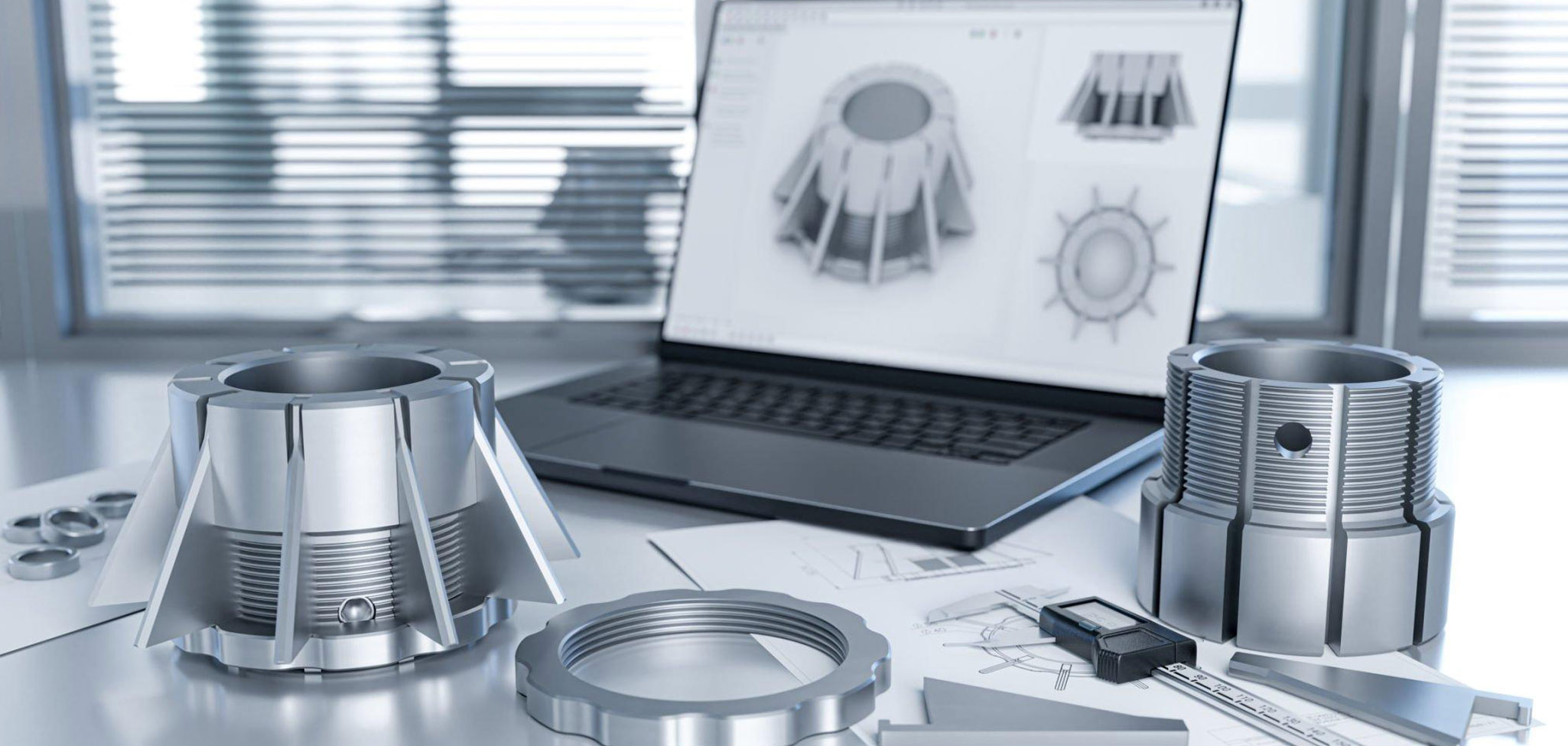 Auf einem Schreibtisch liegen mehrere silberfarbene Maschinenteile und technische Zeichnungen; im Hintergrund steht ein aufgeklappter Laptop, auf dessen Bildschirm ein 3D-CAD-Modell eines ähnlichen Bauteils angezeigt wird.
