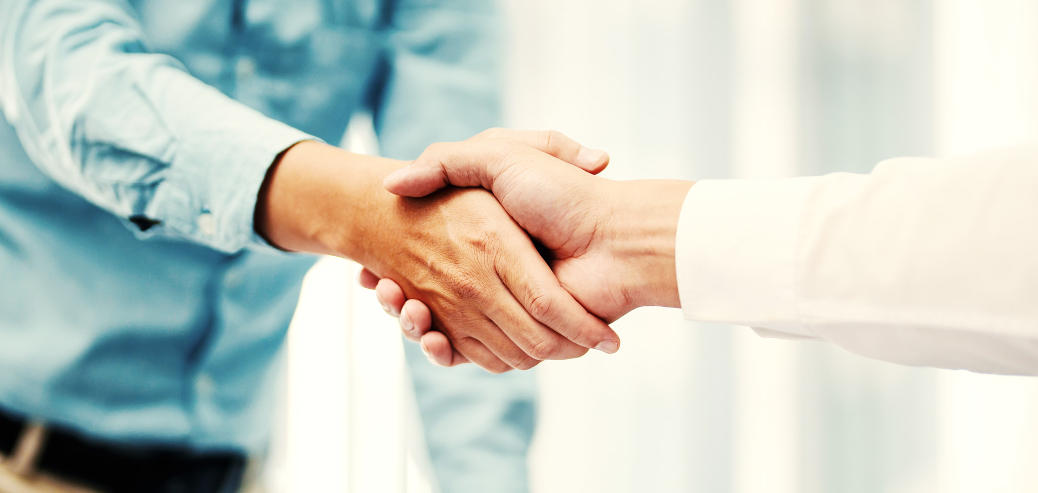 Das Bild zeigt zwei Personen, die sich zur Begrüßung oder als Zeichen einer Einigung die Hand geben; der Fokus liegt auf dem festen Händedruck, wobei von den Personen jeweils nur die untere Hälfte der Arme und die Hände zu sehen sind – eine Person trägt ein weißes Hemd, die andere ein blaues Hemd.