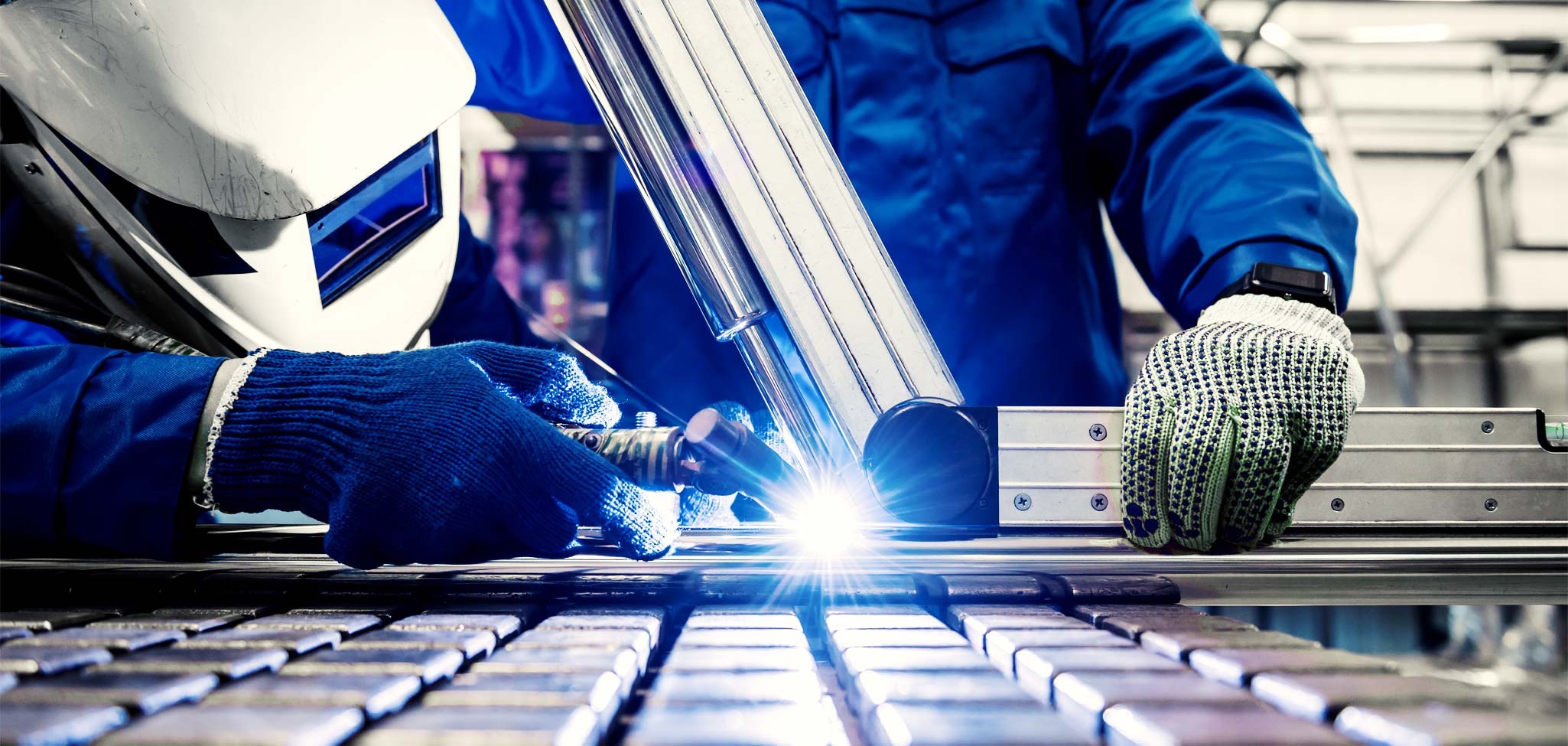 Ein Schweißer in blauer Arbeitskleidung und weißem Schutzhelm führt Schweißarbeiten an einem metallischen Rohrbauteil durch, wobei helle blaue Schweißfunken intensiv aufleuchten. Die Person trägt Schutzhandschuhe und arbeitet in einer industriellen Fertigungsumgebung mit einem Gitterrost im Vordergrund. Die Szene ist aus einer niedrigen Perspektive fotografiert, die den präzisen handwerklichen Arbeitsprozess und das konzentrierte Schweißen in den Fokus rückt.