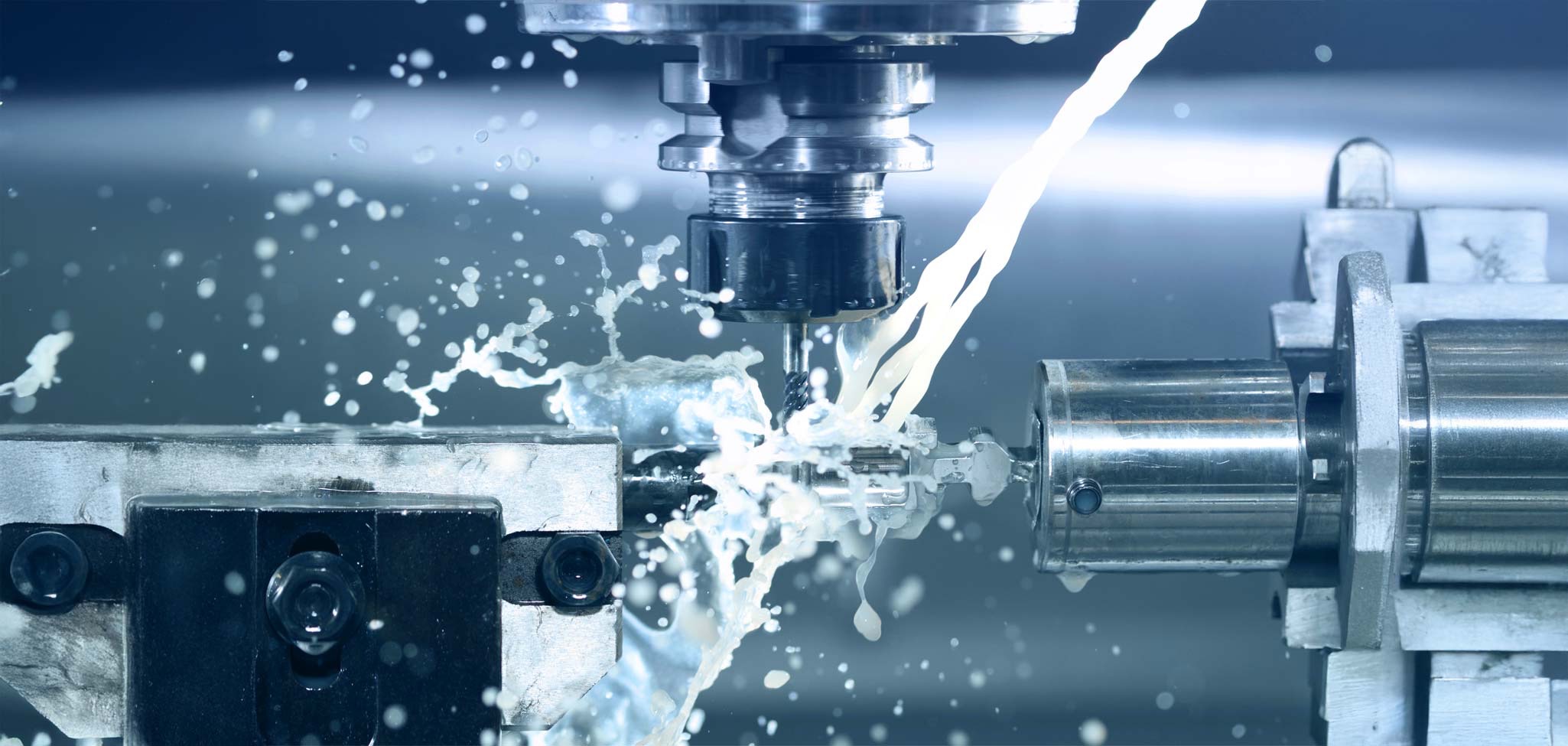 Das Bild zeigt eine Nahaufnahme einer CNC-Fräsmaschine während des Bearbeitungsprozesses: Ein rotierendes Fräswerkzeug bearbeitet ein Metallstück, während Kühlmittel in Strahlen auf das Werkstück und das Werkzeug trifft und dabei zahlreiche Spritzer verursacht. Die Szenerie vermittelt einen modernen, industriellen Eindruck, mit Fokus auf Präzision und Dynamik in der Metallbearbeitung.